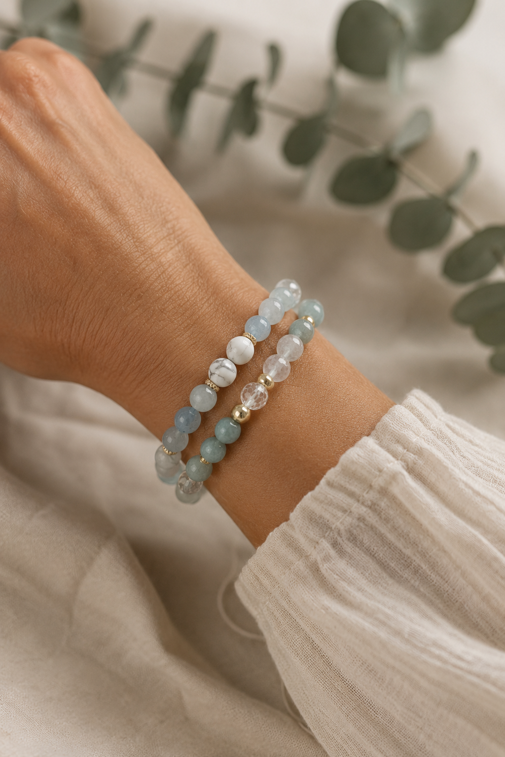 A close-up of a person's wrist wearing two beaded stretch bracelets. One bracelet features light blue and white opaque beads, while the other has translucent pale blue and white beads with a small gold spacer.