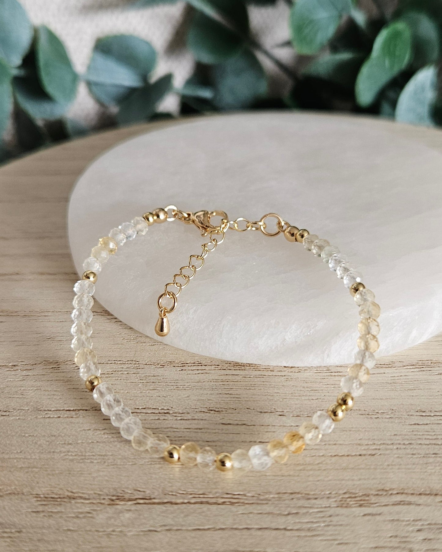 Citrine rondelle bracelet arranged on light wood beside a round selenite plate with eucalyptus in the background
