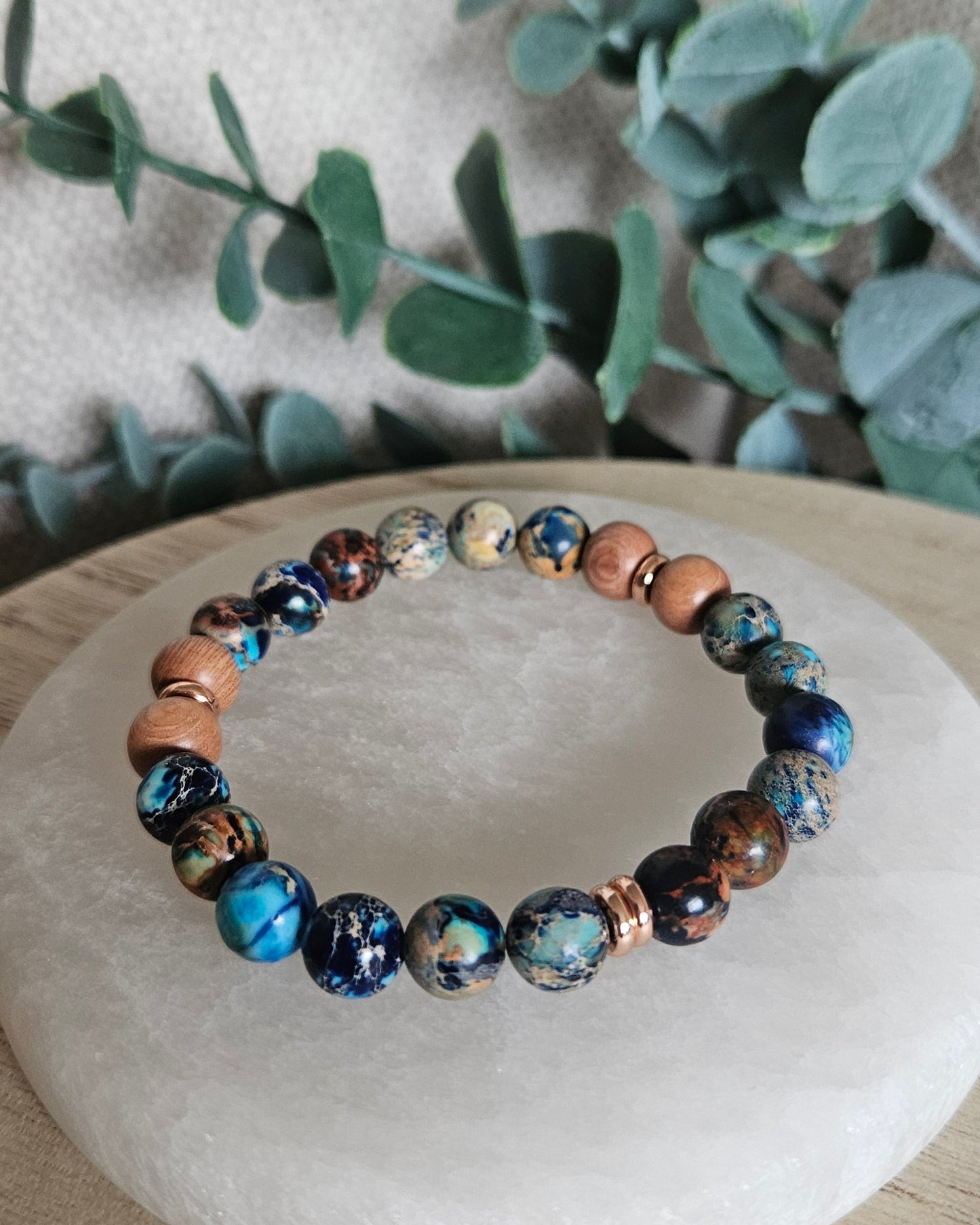 Close-up of multicoloured Ocean Blue Sea Sediment Jasper and Rosewood bracelet beads beside leafy greenery