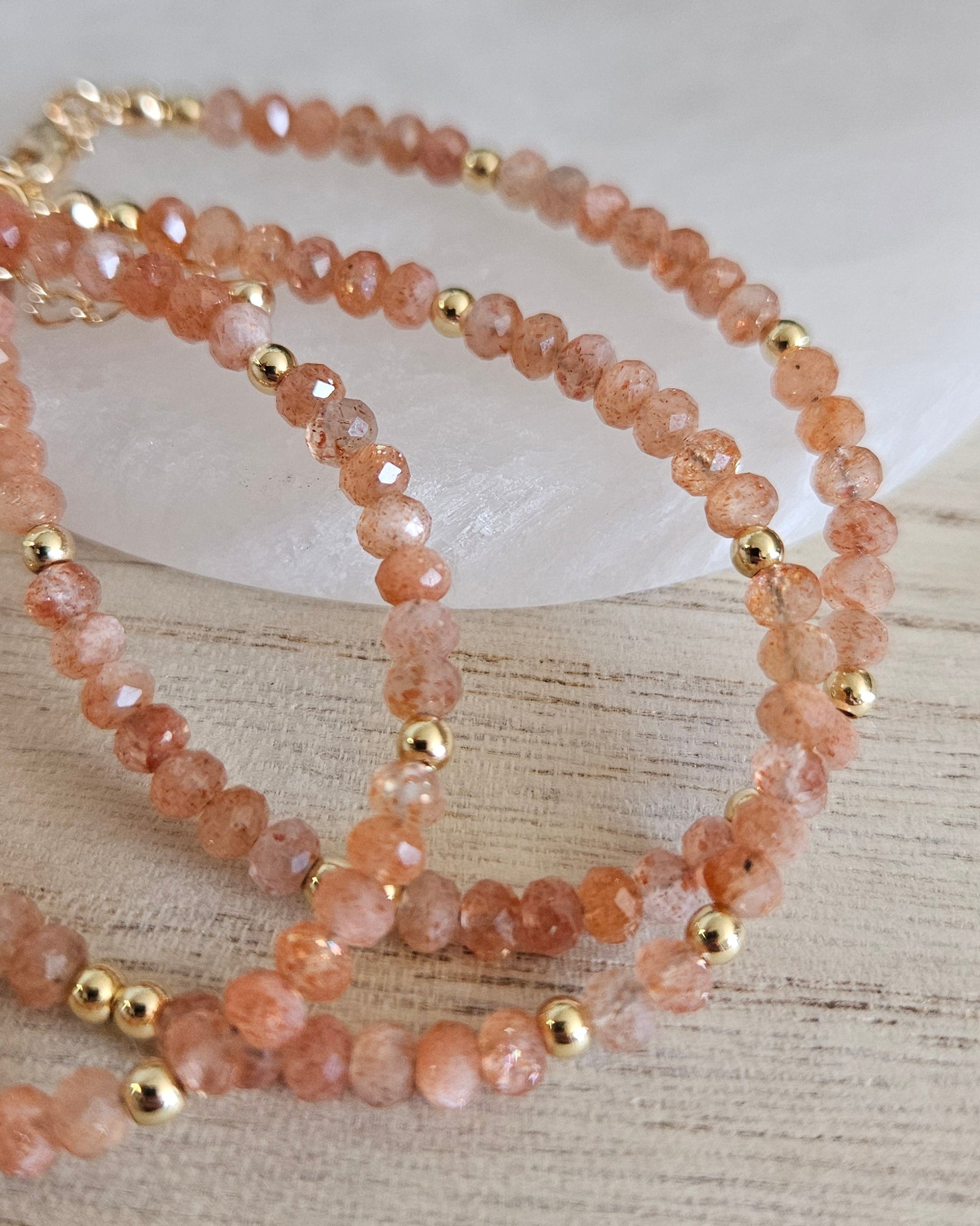 Close-up of several Sunstone bracelets arranged on light wood beside a round selenite plate