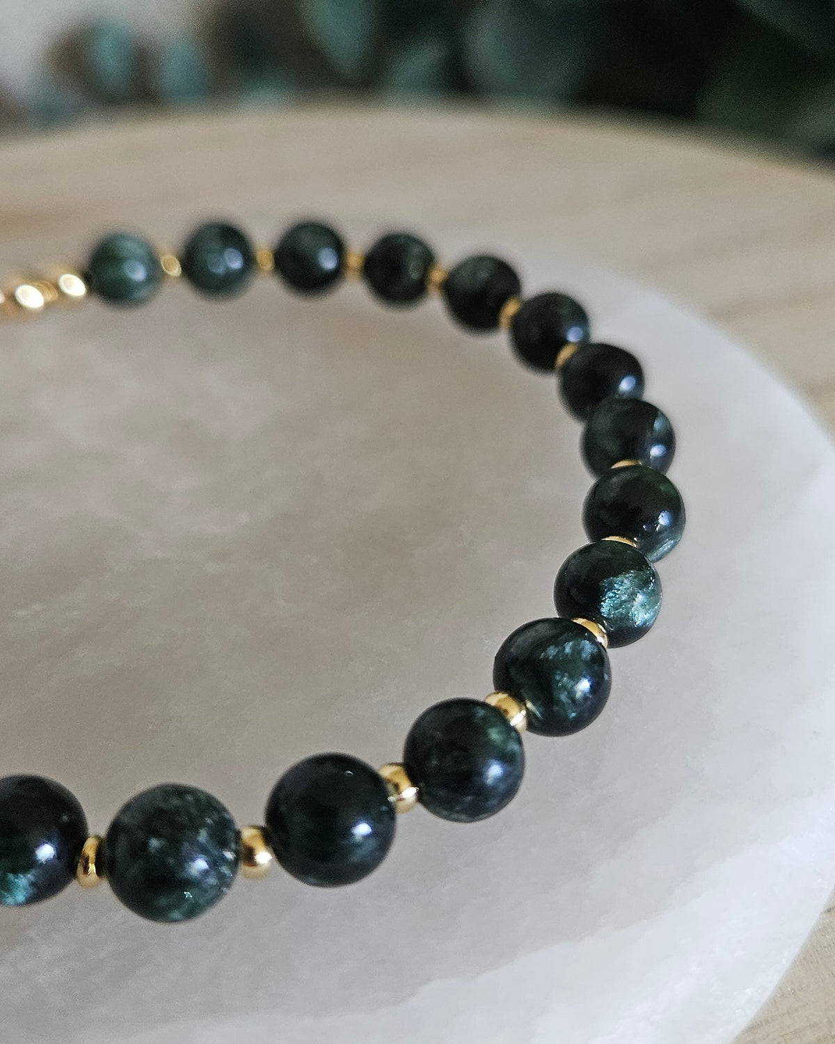 Close view of Seraphinite beads with dark green tones and 14k gold-plated accent beads on a white selenite display