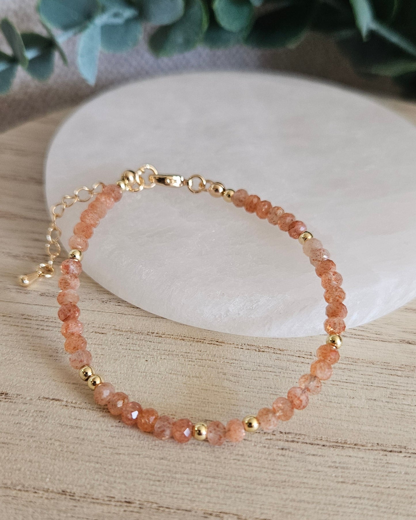 Faceted peach Sunstone bracelet with gold-plated accents, clasp and extender chain displayed beside a selenite plate
