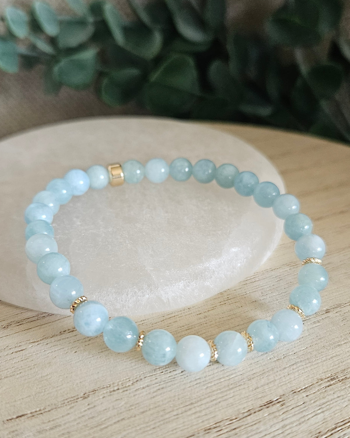 Aquamarine Grace bracelet with light blue gemstone beads and gold-plated accents styled on selenite with greenery behind it
