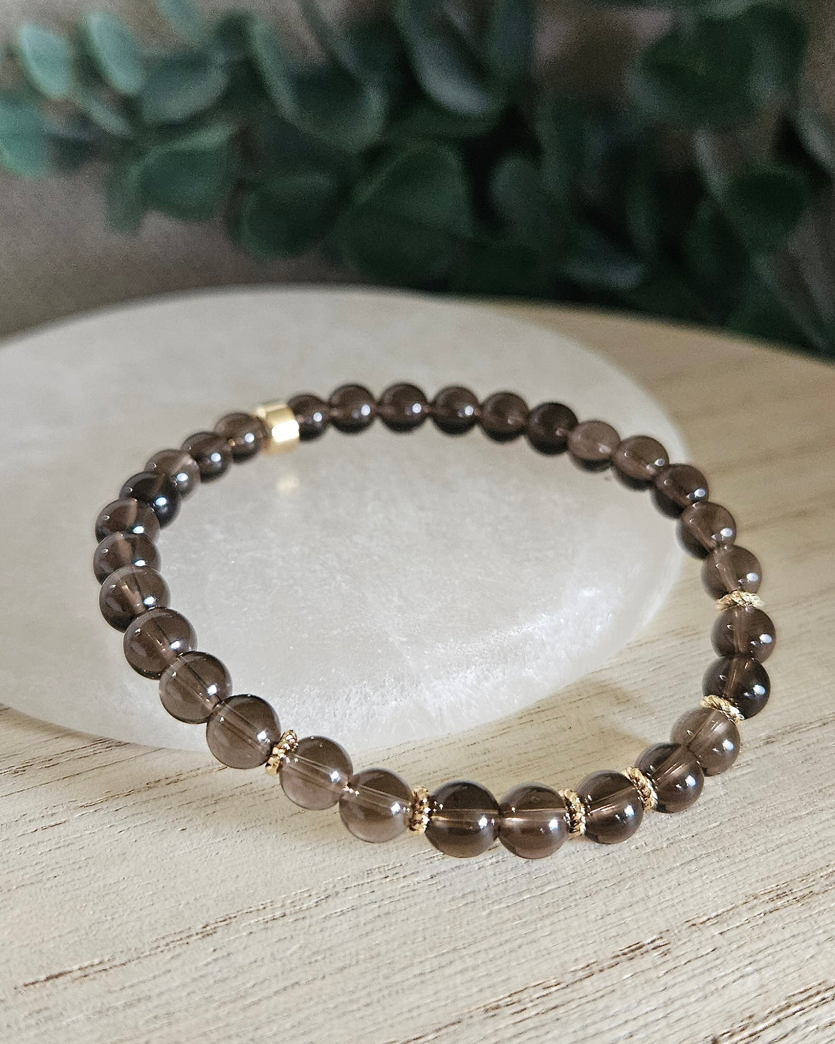 Smoky Quartz Grace bracelet with translucent brown gemstone beads and gold-plated accents styled on selenite with greenery behind it