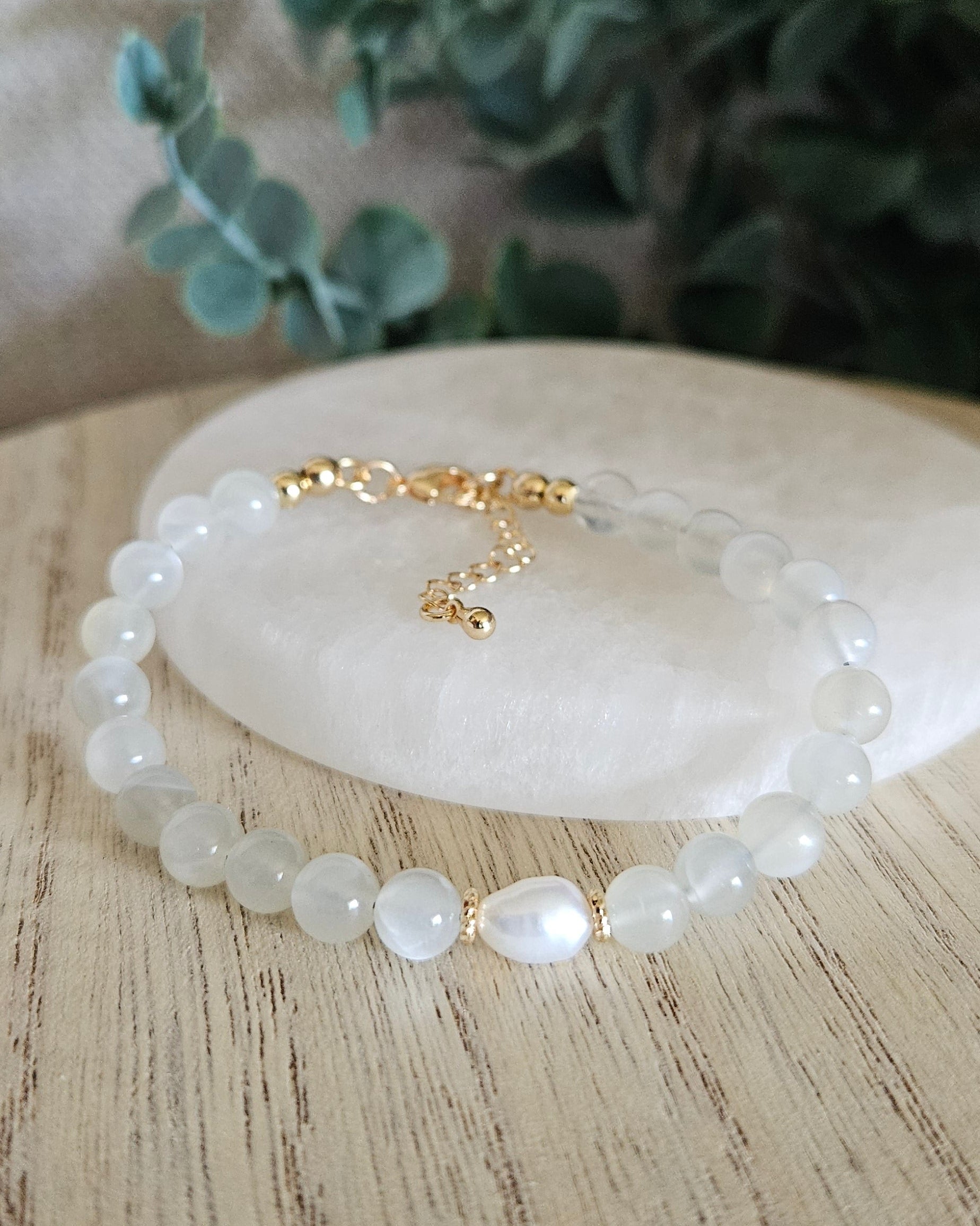 White Moonstone Embrace bracelet arranged before a selenite plate with a freshwater pearl focal and eucalyptus background