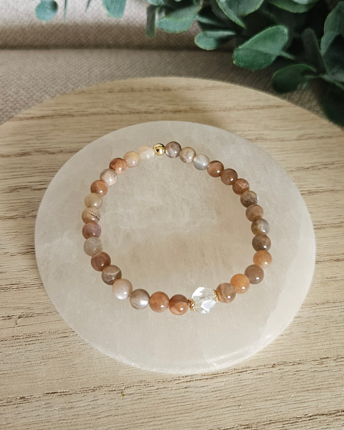 Overhead view of the Chocolate Moonstone Still bracelet with warm neutral beads, Clear Quartz focal and gold accent bead on a white selenite plate