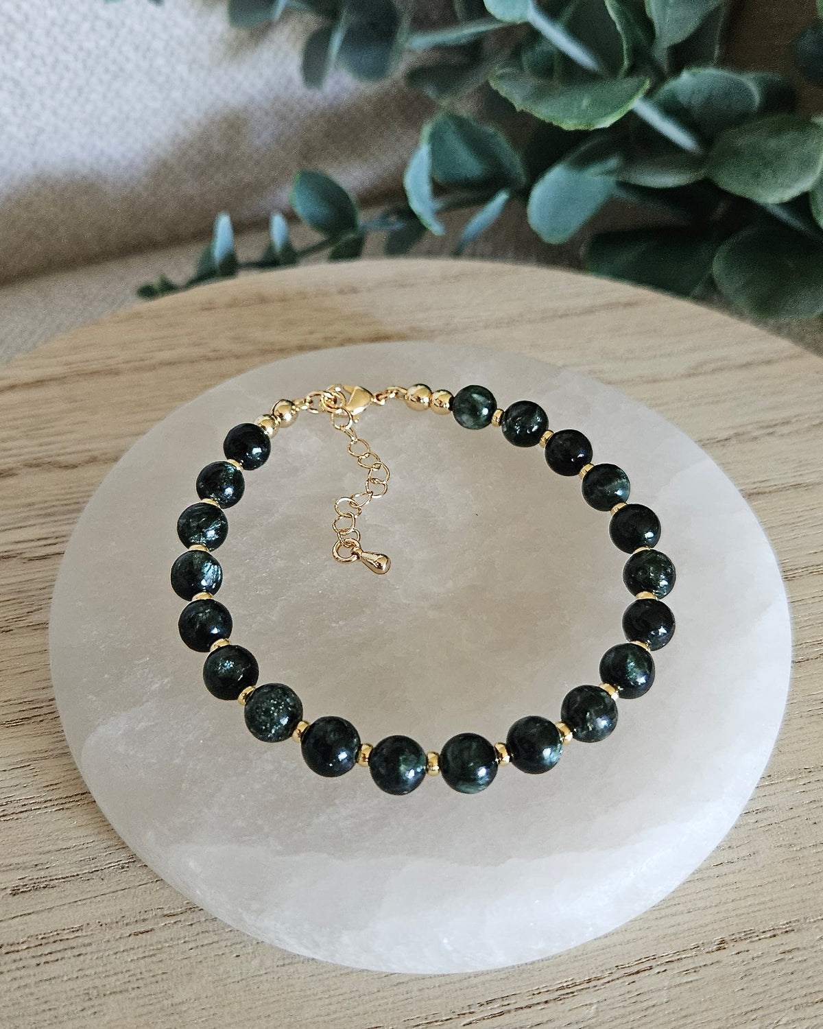 Overhead view of the Seraphinite Echo bracelet with dark green gemstone beads and 14k gold-plated accents shown close up on a white selenite display