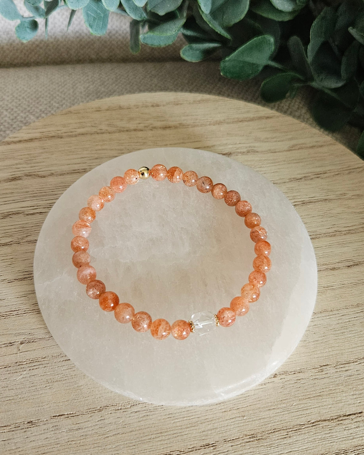Overhead view of the Sunstone Still bracelet with orange 6mm beads, Clear Quartz focal and gold accent bead on a white selenite plate