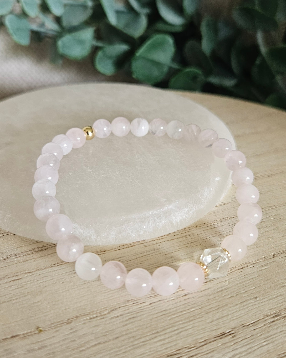 Rose Quartz Still bracelet with 6mm pale pink gemstone beads, Clear Quartz focal and gold accents styled on selenite with greenery behind it
