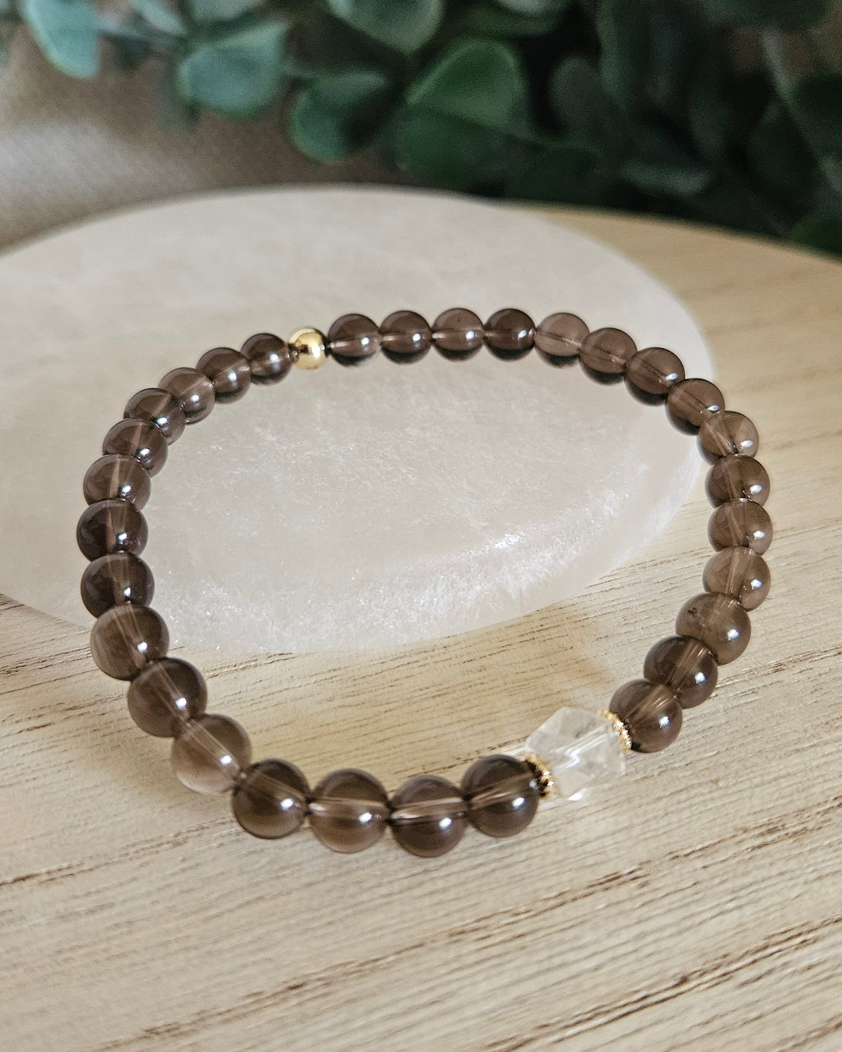 Smoky Quartz Still bracelet with translucent brown gemstone beads, Clear Quartz focal and gold accents styled on selenite with greenery behind it