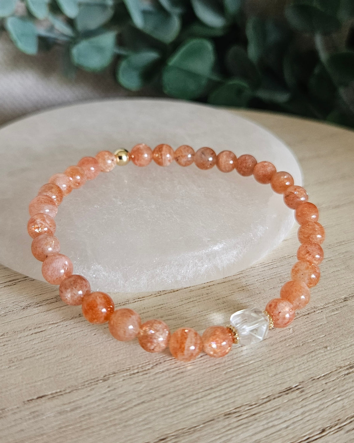 Sunstone Still bracelet with 6mm orange gemstone beads, Clear Quartz focal and gold accents styled on selenite with greenery behind it

