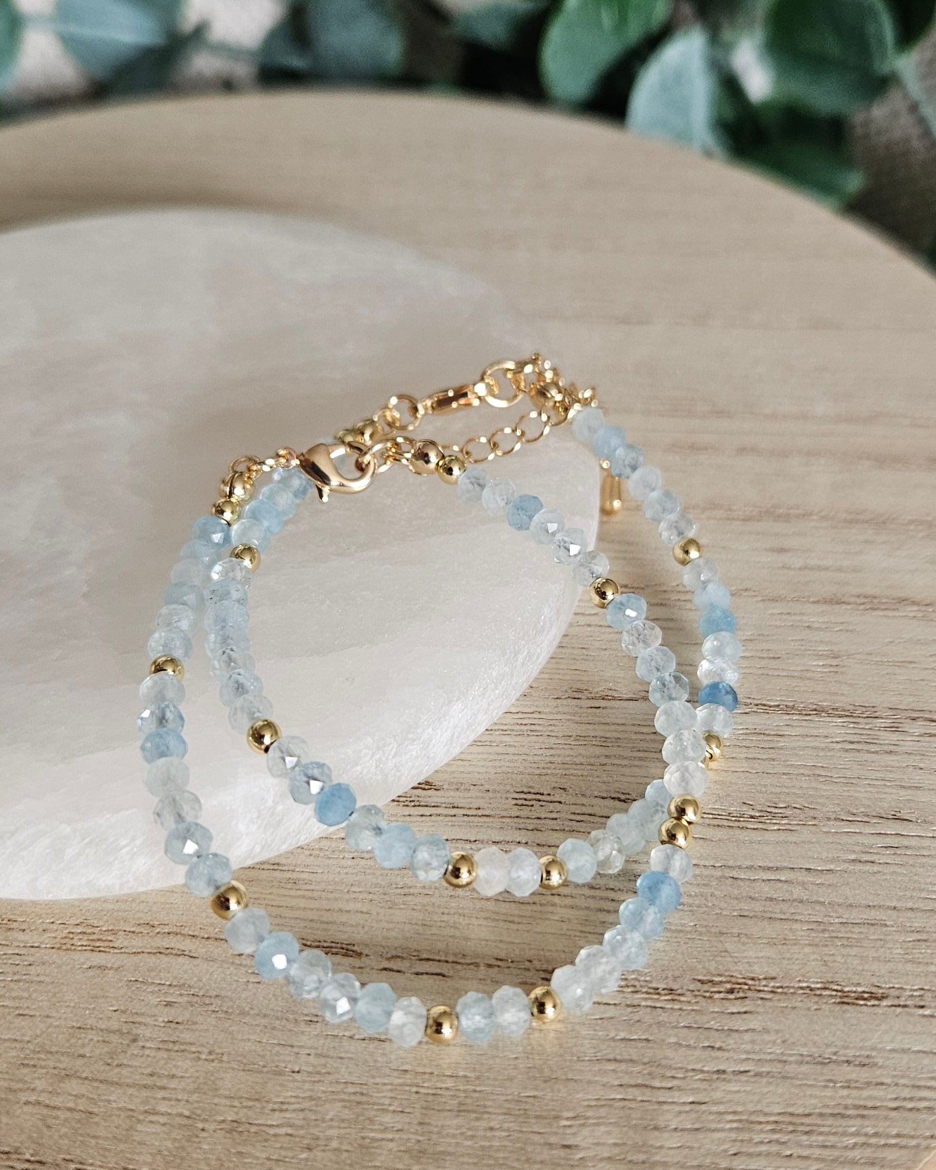 Two layered Aquamarine bracelets with gold-plated clasps and extender chains displayed beside selenite